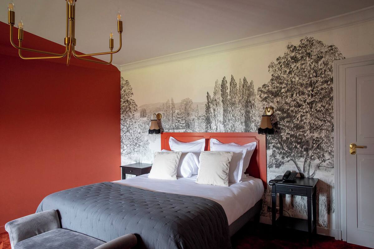 Een slaapkamer met een bed, nachtkastje, muurschildering en een kroonluchter.