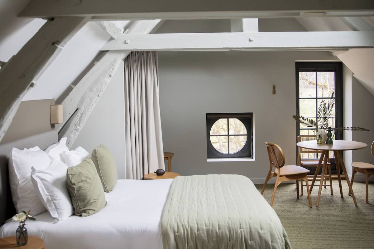 Een slaapkamer met een schuine kap, een bed met witte lakens, een ronde tafel met stoelen en een raam met uitzicht op bomen.