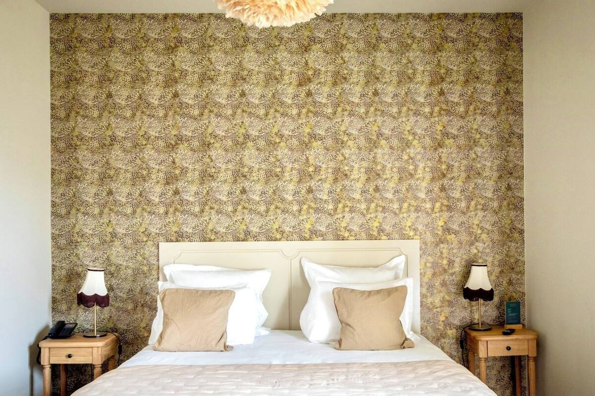 Een hotelkamer met behang met een patroon, een groot bed met twee kussens en twee nachtkastjes met lampen.