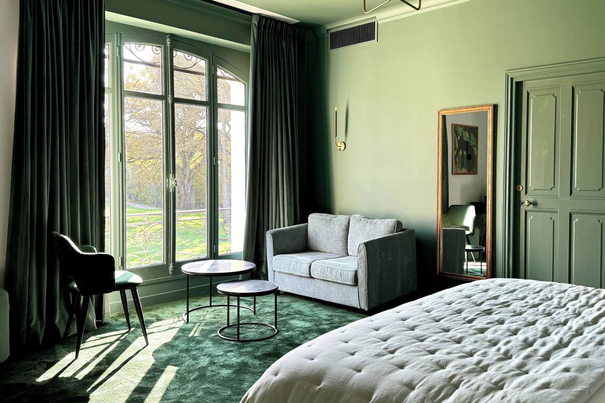 Hotelkamer met een groot bed, een grijze bank, een klein rond tafeltje en een stoel. Er is een groot raam met gordijnen, een deur en een kroonluchter.