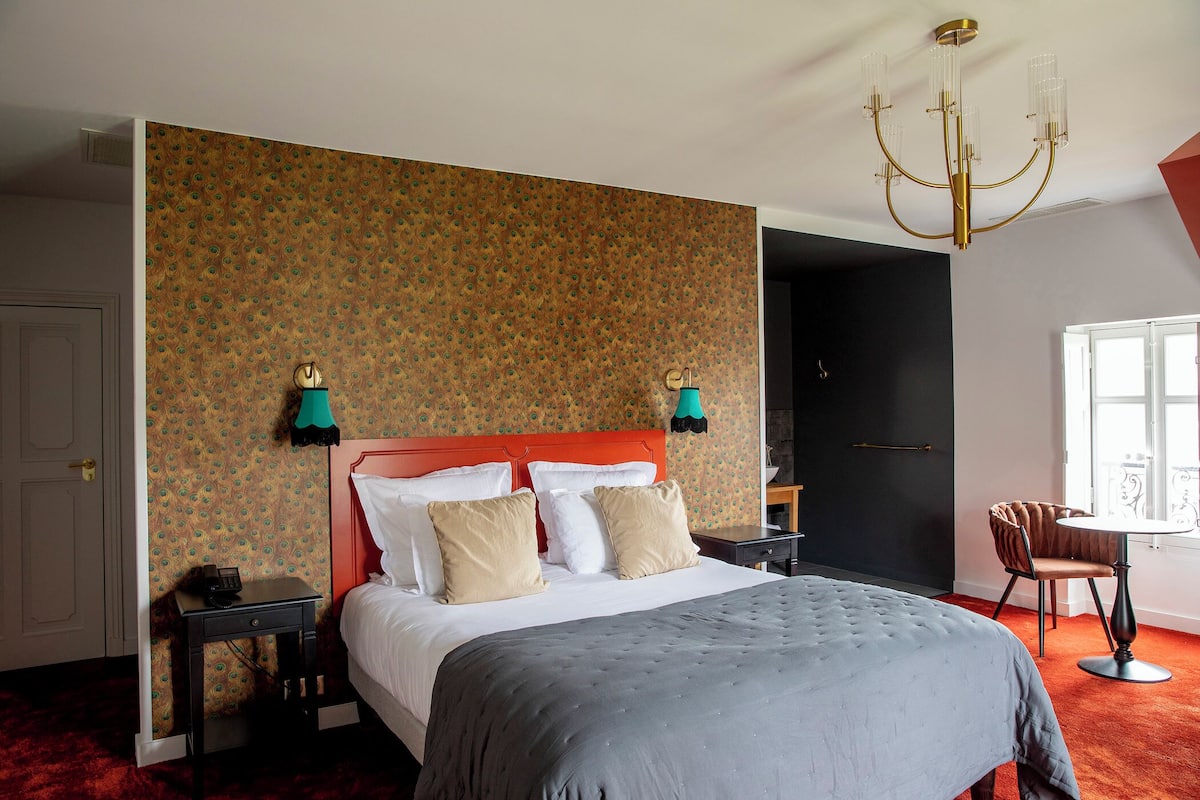 Hotelkamer met een groot bed, een rood hoofdeinde, behang met een patroon, een kroonluchter, een bureau met een lamp en een raam met uitzicht.