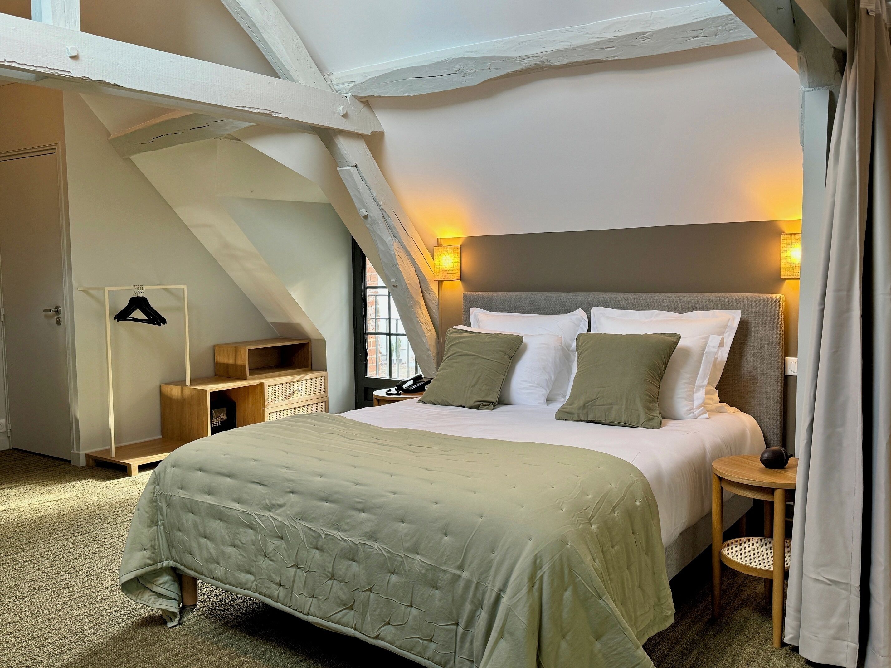 A bedroom with a lofted ceiling, a bed with a green bedspread, a bedside table with a lamp, and a hanging coat rack.