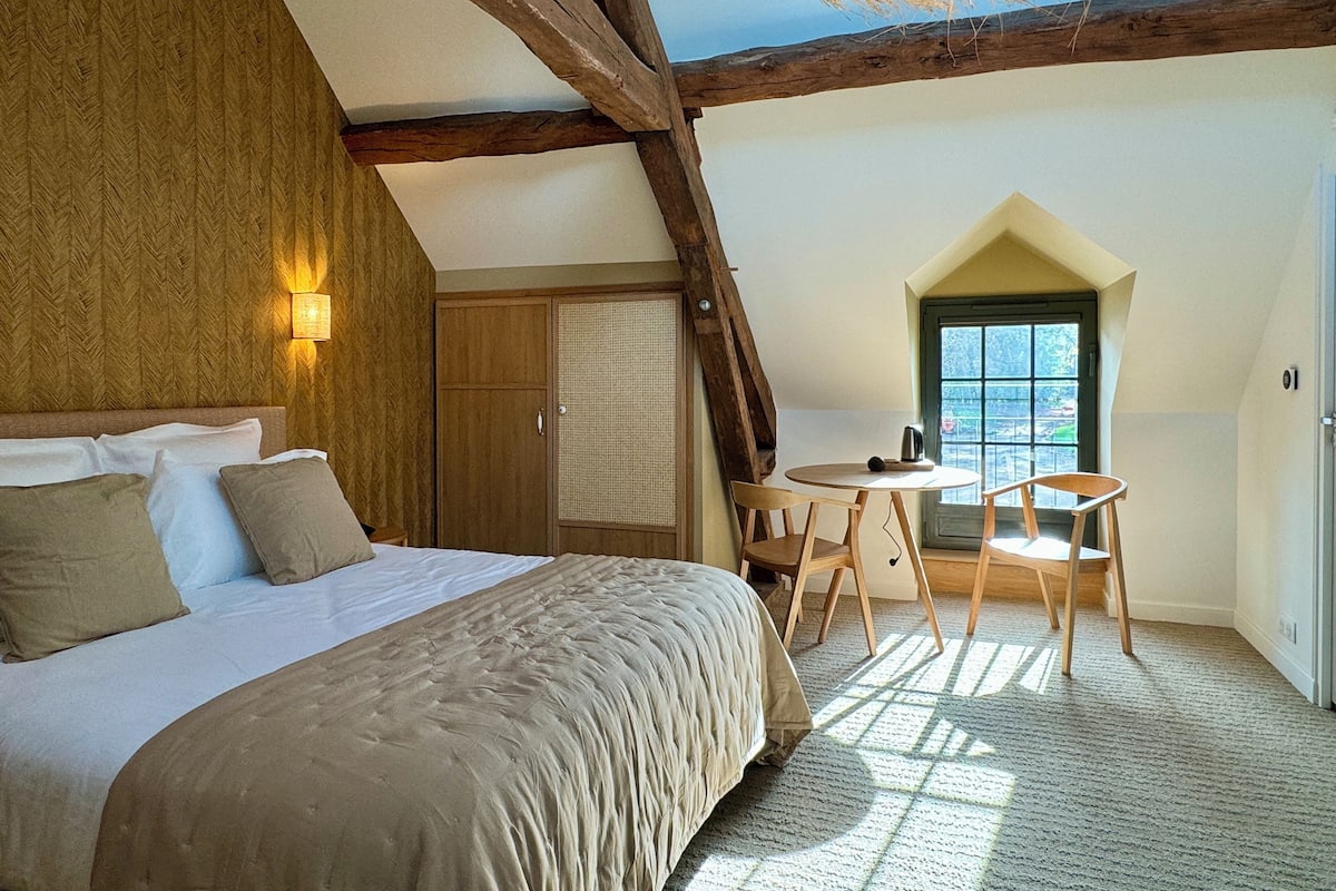 Een slaapkamer met een bed, houten plafond, een raam en een kleine tafel met stoelen.