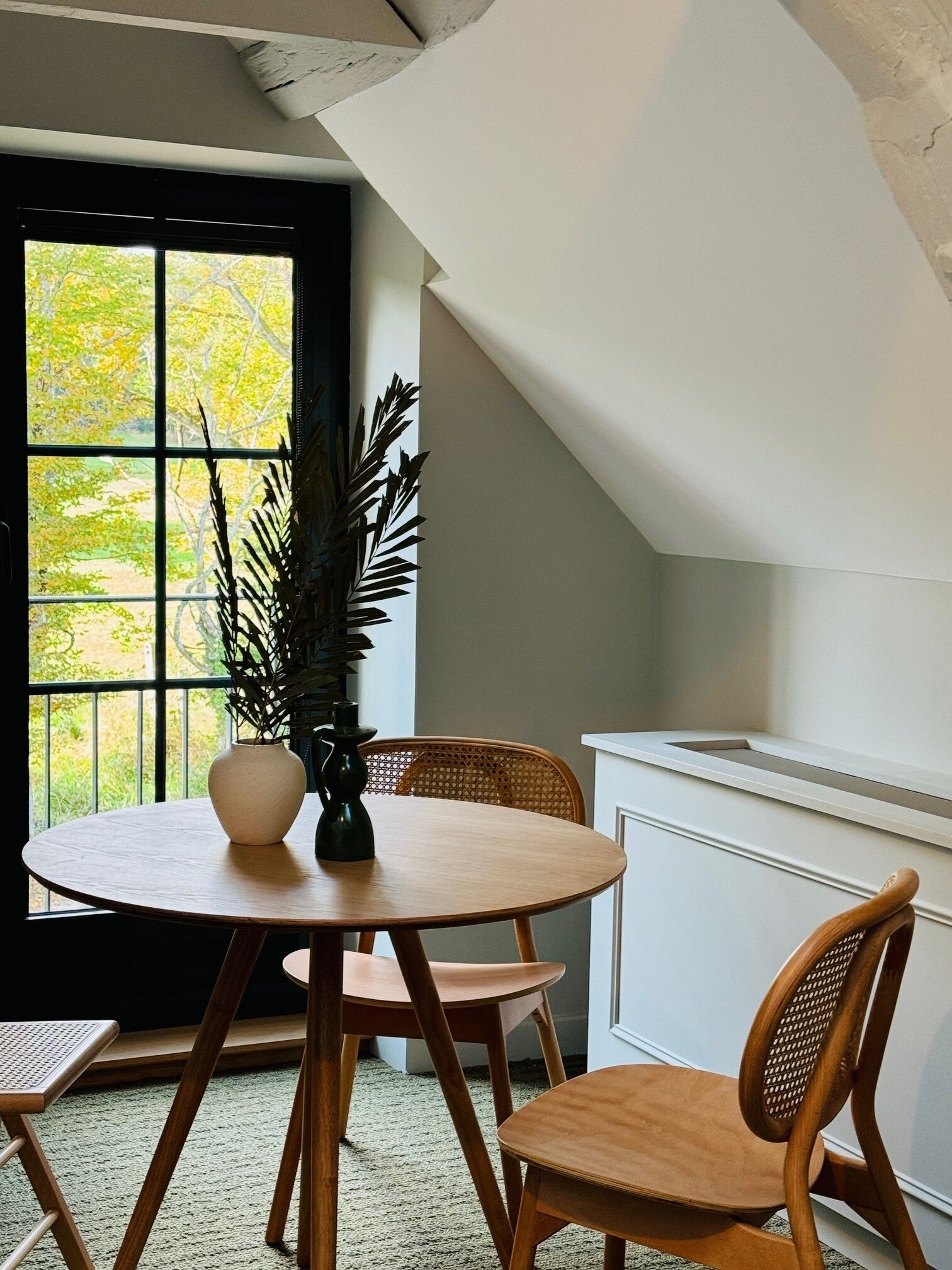 Un comedor con una mesa redonda de madera, dos sillas de madera, un jarrón con una planta y vistas a través de una ventana con marco negro.
