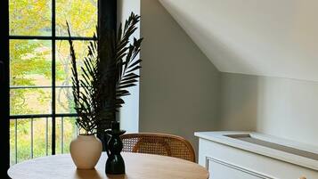 Un comedor con una mesa redonda de madera, dos sillas de madera, un jarrón con una planta y vistas a través de una ventana con marco negro.