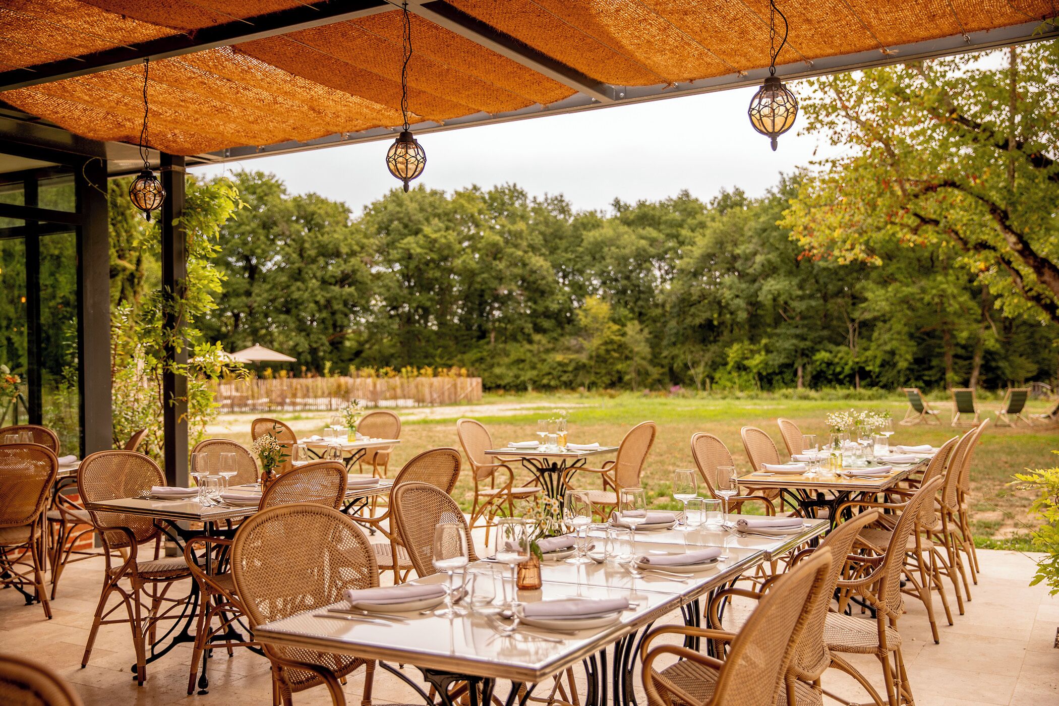 Un espacio de comedor al aire libre con sillas y mesas de mimbre, techo de paja y vistas a un jardín y árboles.