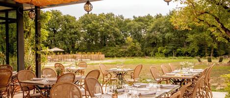 Un espacio de comedor al aire libre con sillas y mesas de mimbre, techo de paja y vistas a un jardín y árboles.