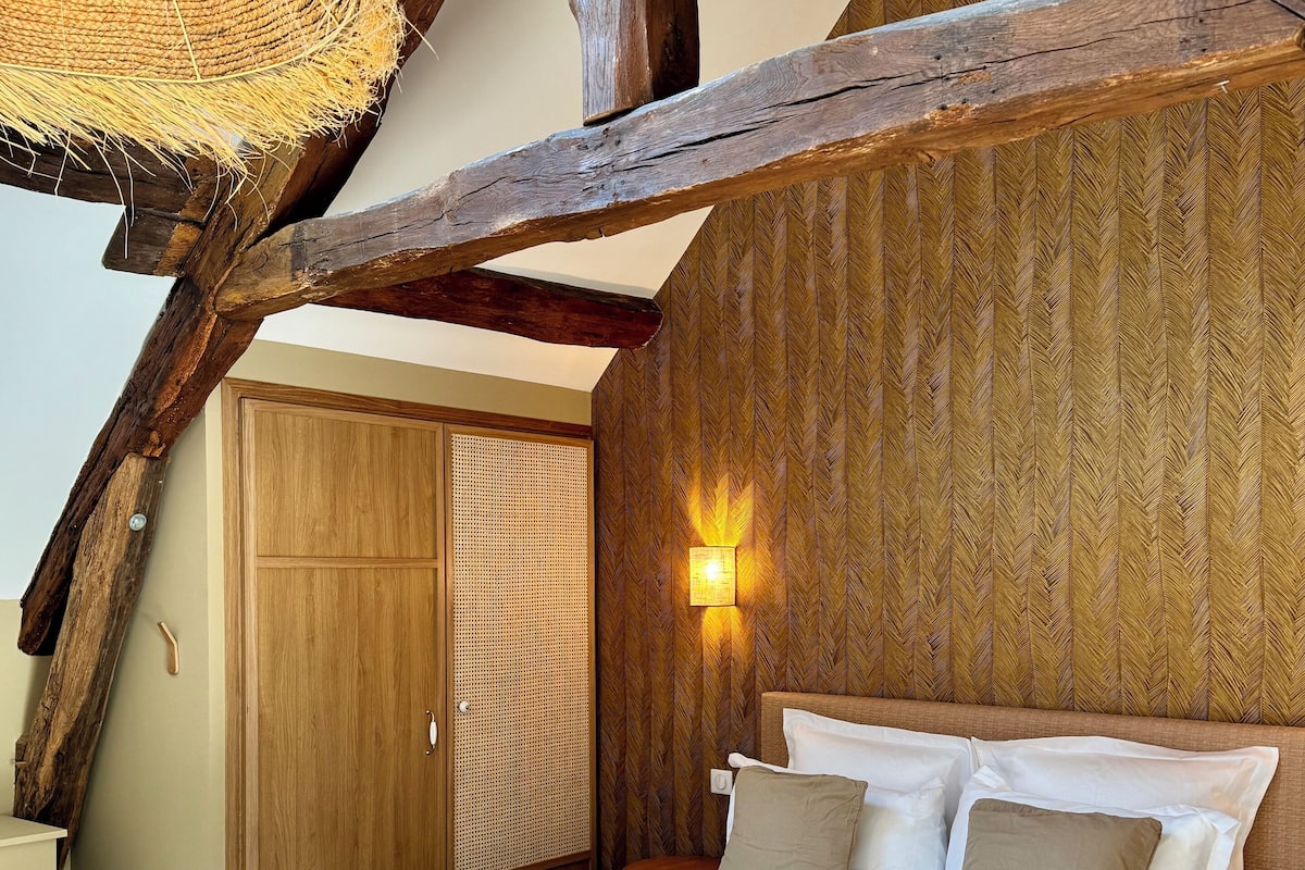 Een slaapkamer met een bed, houten balken, een geweven lamp en een wand met een herringbone-patroon, plus een nachtkastje.