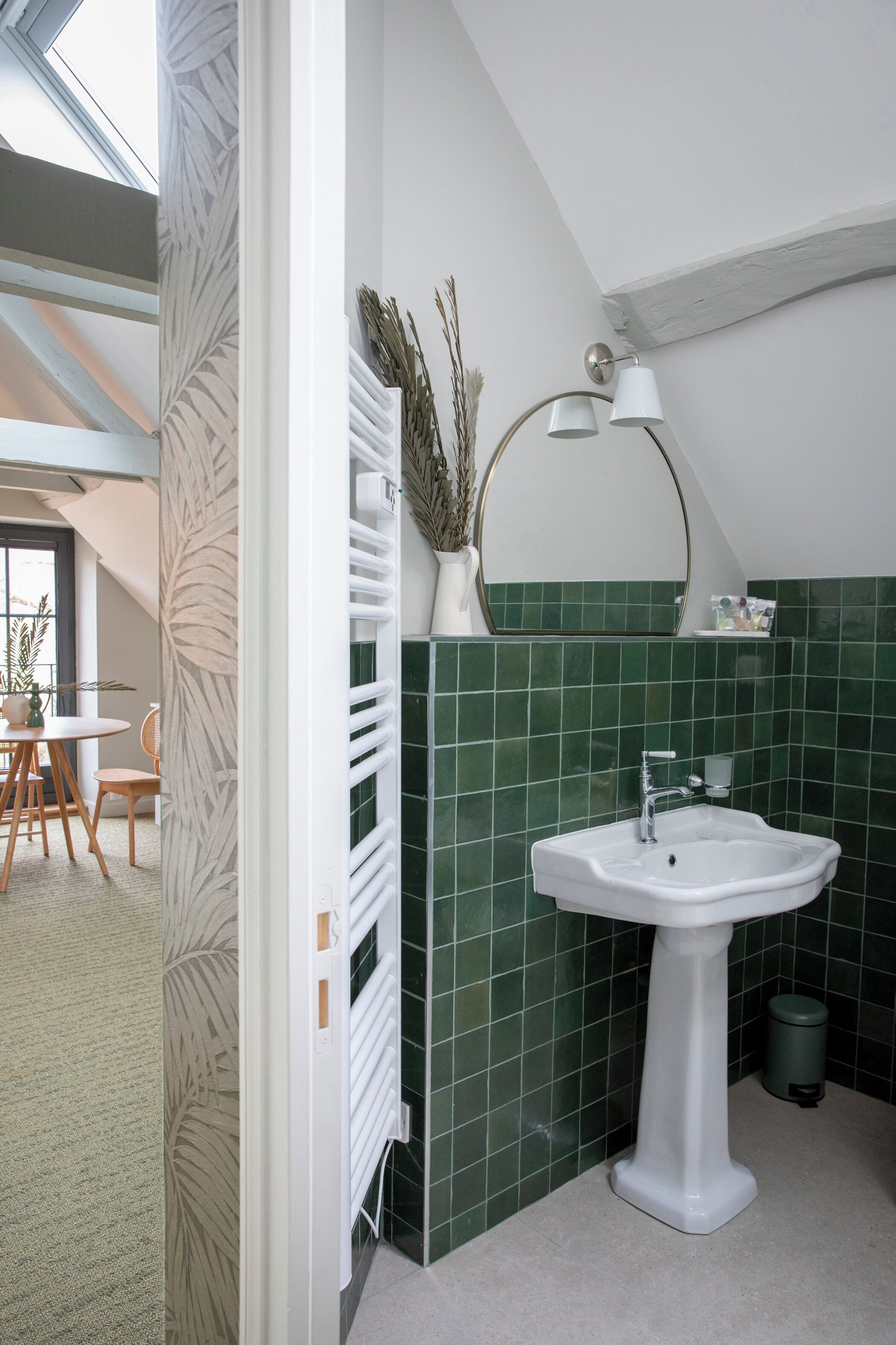 Un baño con lavamanos pedestal blanco, azulejos verdes, radiador blanco, un espejo y una lámpara.