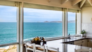 A kitchen with large windows overlooking the sea, a dining area with chairs, and a view of distant land. - Absolute Ocean Frontage 1 The Strand Pt Elliot (Port Elliot)