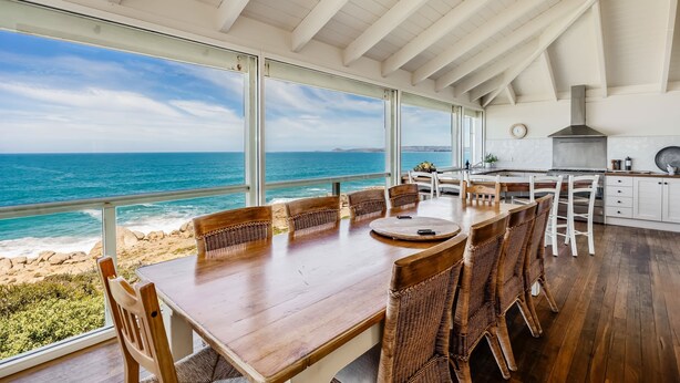 A dining area with a view of the ocean, wooden furniture, and a kitchen with white cabinets. - Absolute Ocean Frontage 1 The Strand Pt Elliot (Port Elliot)