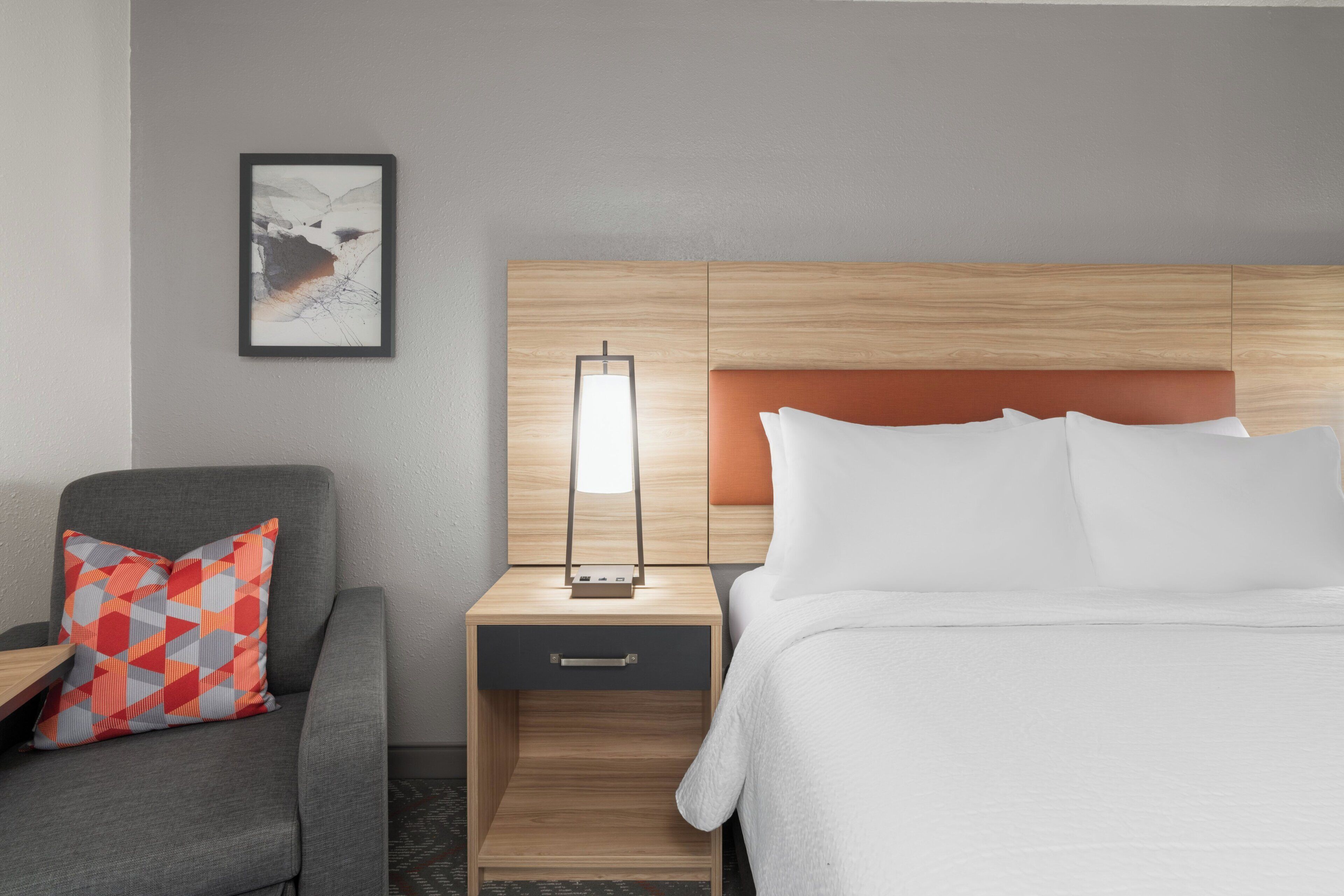 A hotel room with a bed, a nightstand with a lamp, a chair with a patterned pillow, and a framed picture on the wall.
