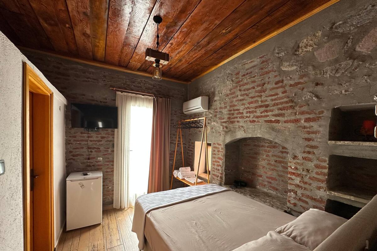 Una habitación rústica con paredes de ladrillo visto, techo de madera, cama con colcha color beige, refrigerador blanco pequeño, perchero colgante y chimenea de ladrillo.