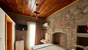 A rustic room with a brick wall, a wooden ceiling, a bed, a small table, and a hanging clothes rack. - Moj Dom Küçükköy (Ayvalik)