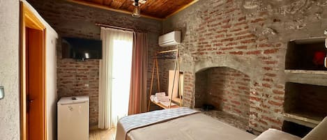 A rustic room with a brick wall, a wooden ceiling, a bed, a small table, and a hanging clothes rack.