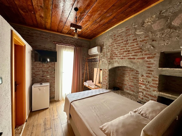 Um quarto rústico com parede de tijolos, teto de madeira, uma cama, uma mesinha e um cabideiro.