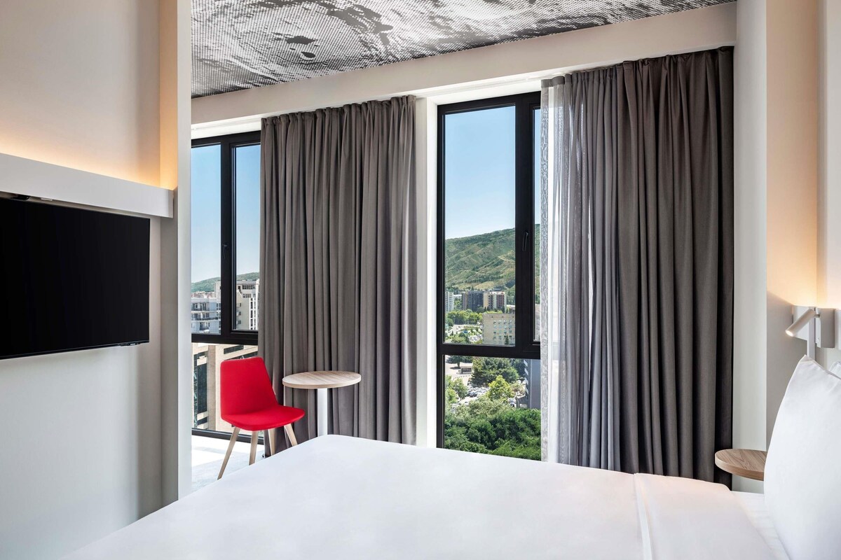 Ein Hotelzimmer mit einem Bett, einem Fernseher, einem roten Sessel, einem kleinen runden Tisch und großen Fenstern mit Vorhängen.