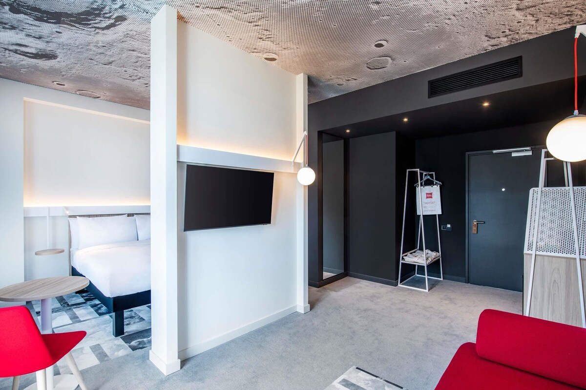 Habitación de hotel moderna con una televisión de pantalla plana montada en la pared, una pequeña mesa redonda, una silla roja y un sofá rojo.
