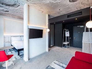 Habitación de hotel moderna con una televisión de pantalla plana montada en la pared, una pequeña mesa redonda, una silla roja y un sofá rojo.