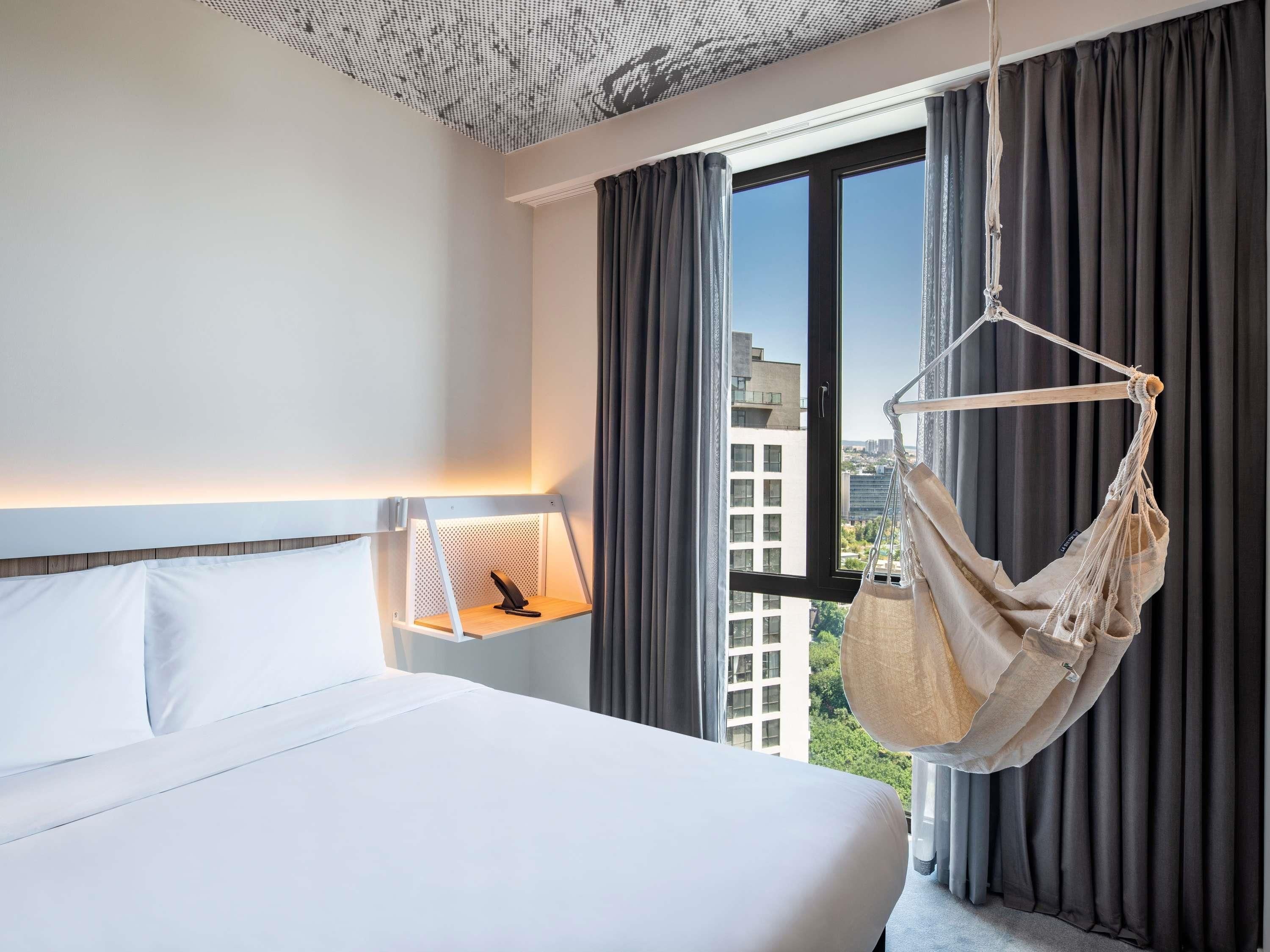 A hotel room with a bed, a hanging chair, a nightstand with a lamp, and a window with curtains.