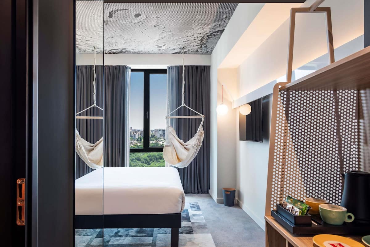 Quarto de hotel moderno com uma cama grande, vista para a cidade, televisão e mesa com cafeteira.