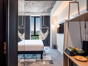 A modern hotel room with a large bed, a view of the city, a TV, and a desk with a coffee machine.