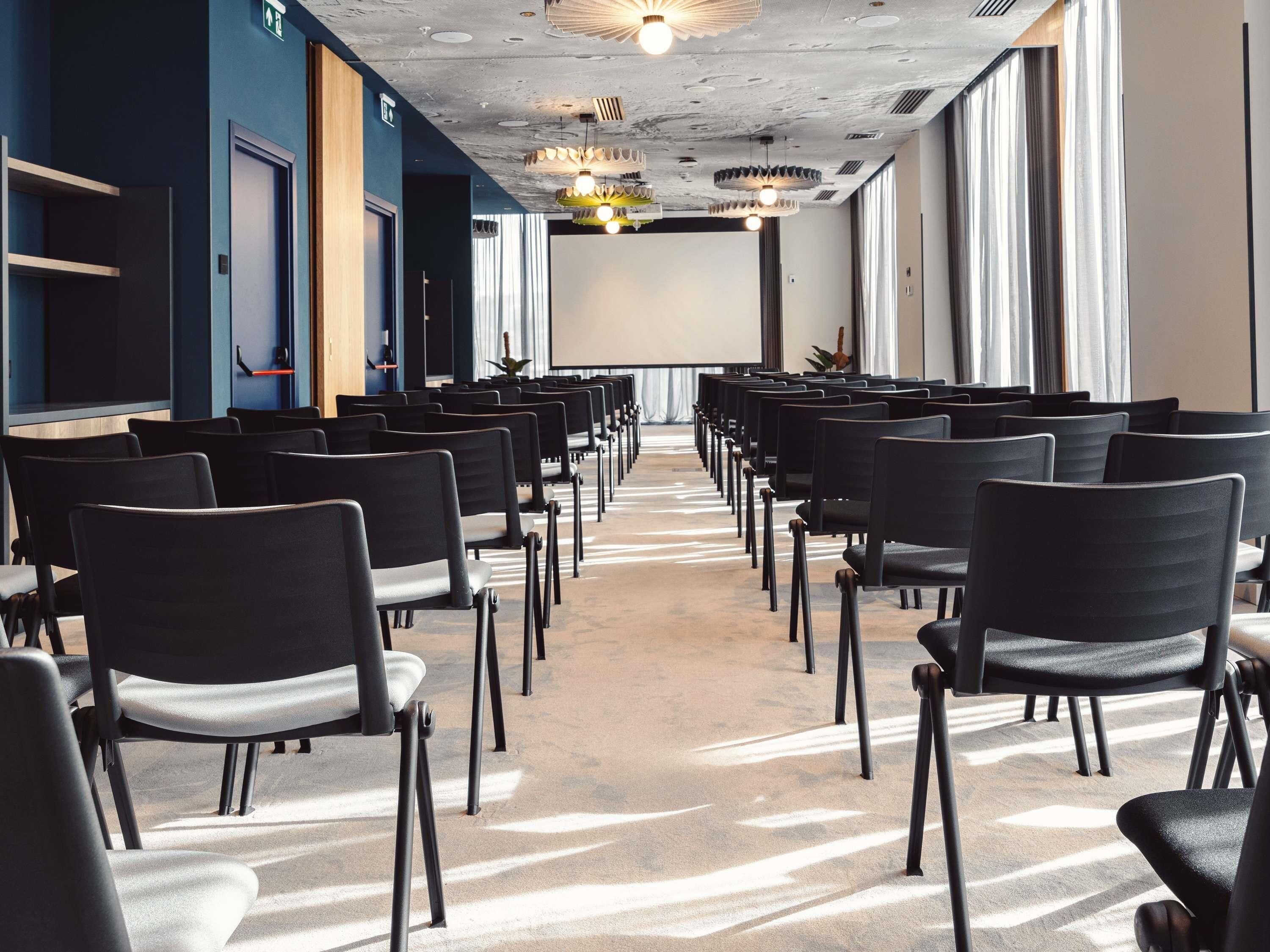 Une salle de conférence avec des rangées de chaises, un écran de projection et une moquette à motifs.