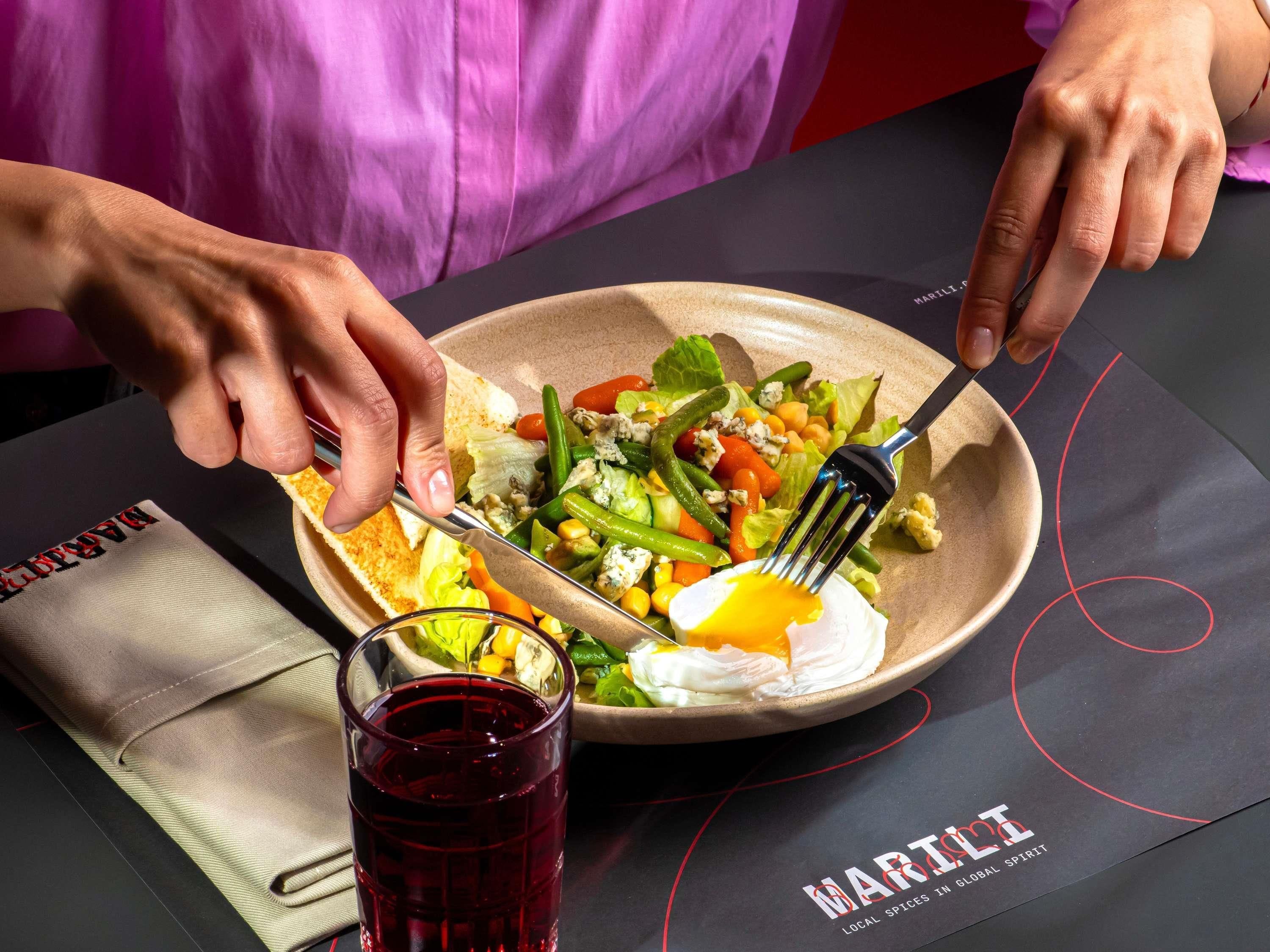 A person cutting a salad with a fork and knife on a plate, with a glass of red drink beside it.