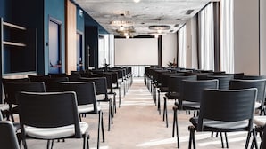 Un salón de conferencias con filas de sillas, una pantalla de proyector e iluminación de techo original.
