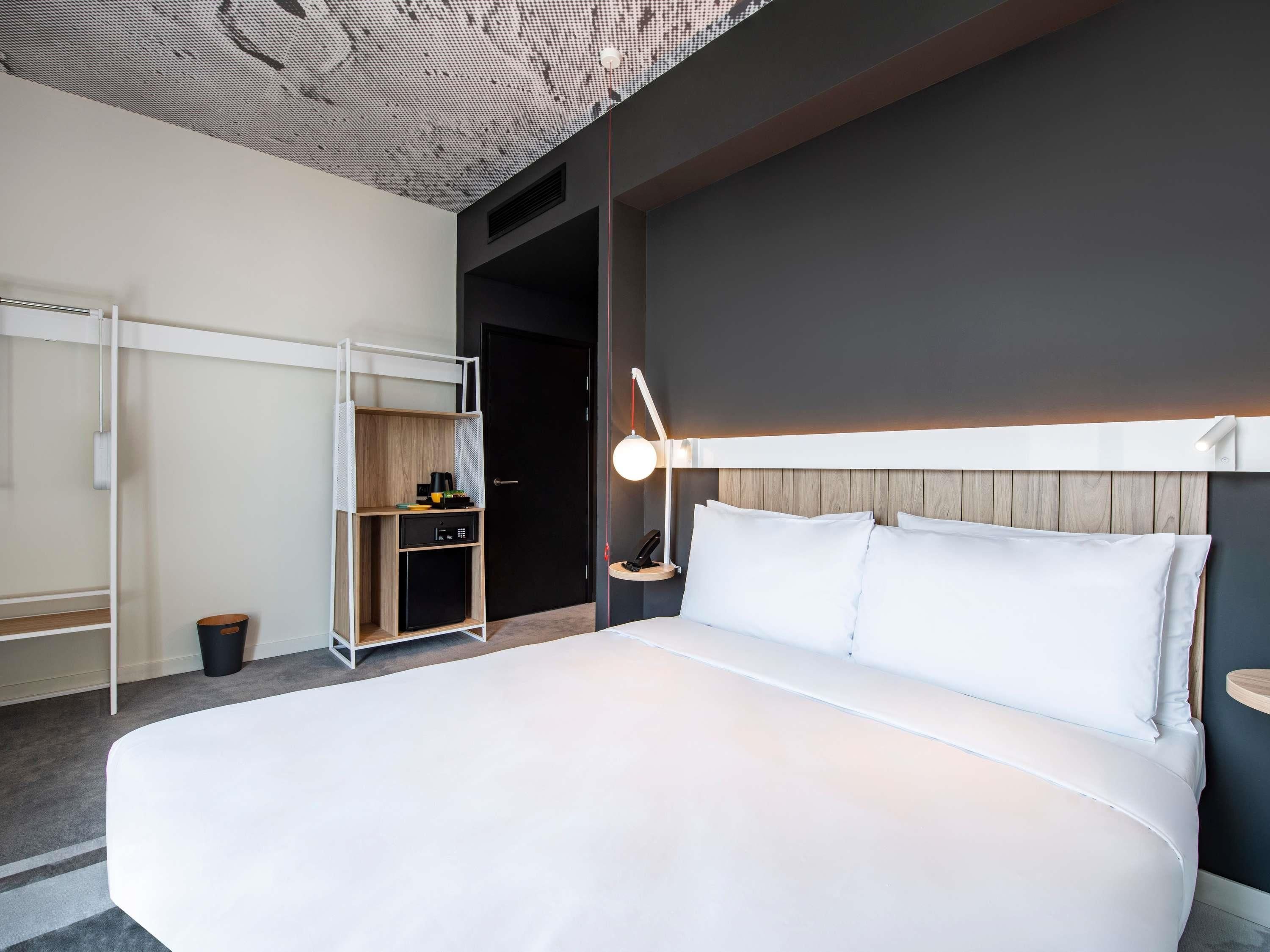 Ein modernes Hotelzimmer mit einem großen Bett, einem Schreibtisch mit Stuhl, einer kleinen Küchenzeile und einer Decke mit einem einzigartigen Muster.