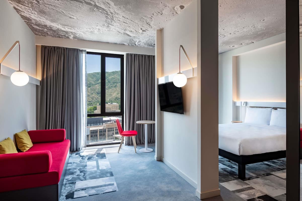 Quarto de hotel com sofá vermelho, uma cama, televisão e varanda com vista para as colinas.