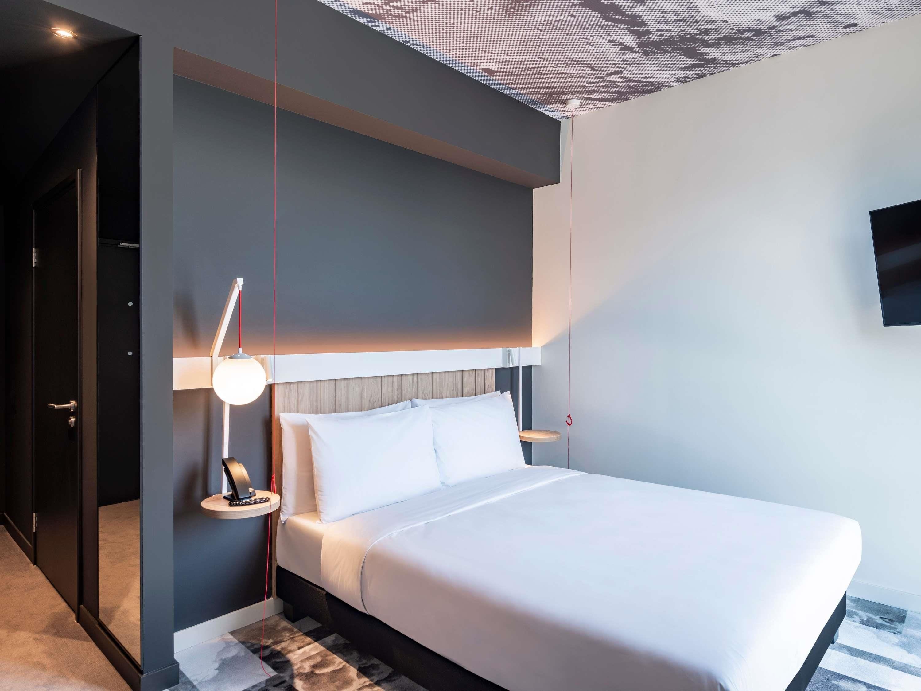 A modern hotel room with a large bed, a bedside table with a lamp, a wall-mounted TV, and a unique ceiling design.
