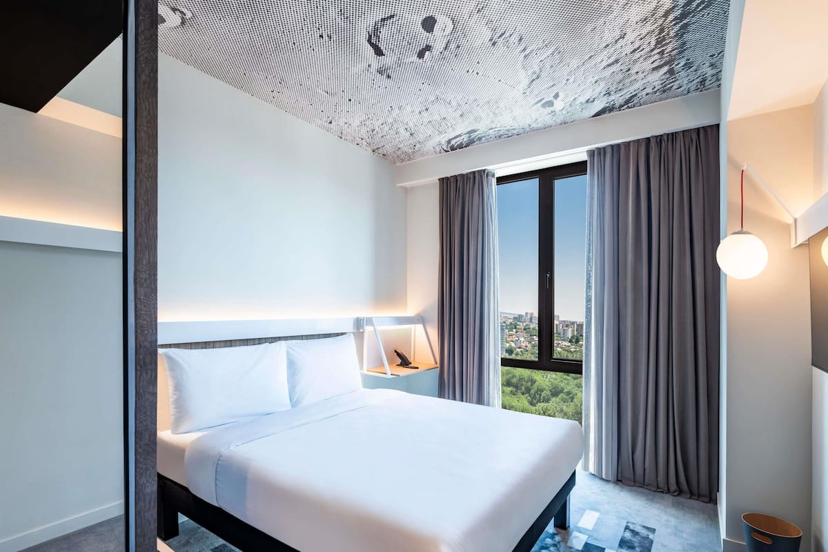 Quarto de hotel moderno com uma cama grande, vista da cidade pela janela e teto com padrão único.