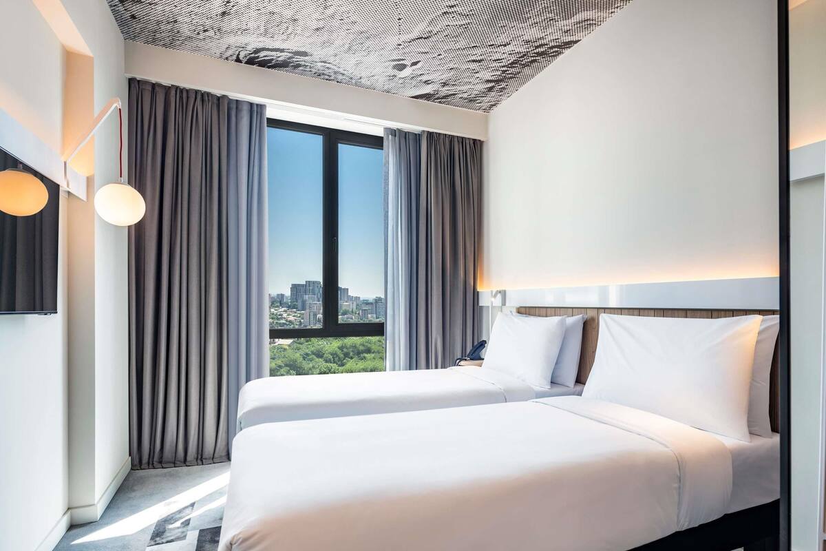 Quarto de hotel moderno com duas camas, vista para a cidade e teto com padrão.