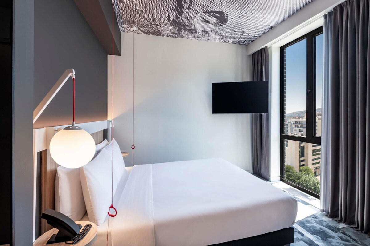 A modern hotel room with a bed, a desk with a lamp, a flat-screen TV, and a large window offering a city view.