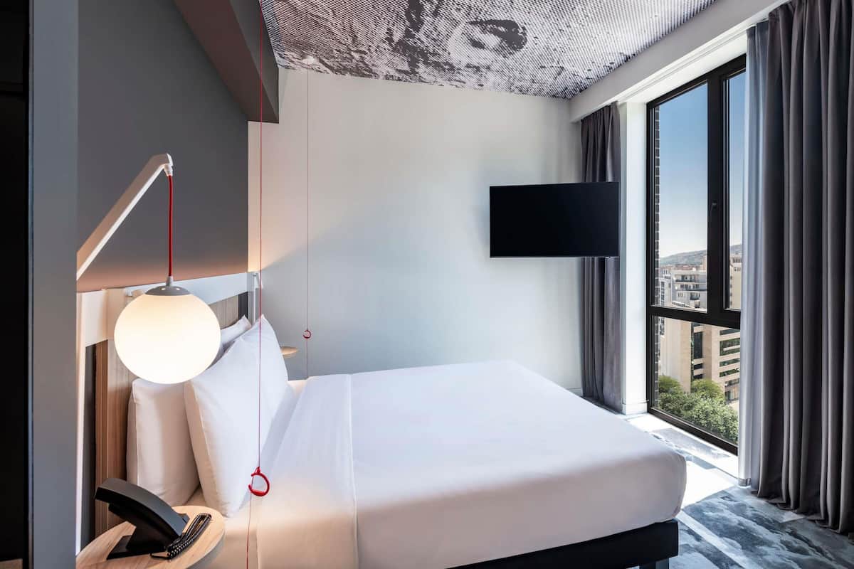 Quarto de hotel moderno com cama, escrivaninha com luminária, TV de tela plana e uma grande janela com vista para a cidade.