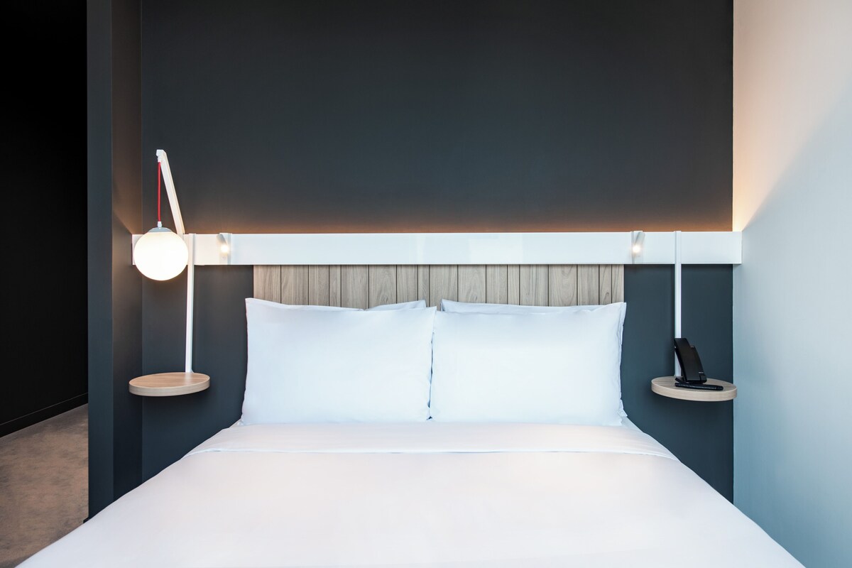 Una habitación de hotel moderna con una cama grande, mesitas de noche y una lámpara fijada a la pared.