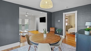 A modern living room with a round wooden table, two chairs, a potted plant, and a television on the wall. - Renovated Water Haus ⎥ +8 beds ⎥ Historic Waterfront

 (Warren)