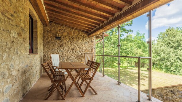 A covered patio with a stone wall, wooden table and chairs, and a view of greenery. - Le Casaline Di Sopra Near Massa (Massa Marittima)