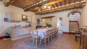 A spacious kitchen with wooden beams, a dining table with chairs, and a staircase leading to another level. - Le Casaline Di Sopra Near Massa (Massa Marittima)