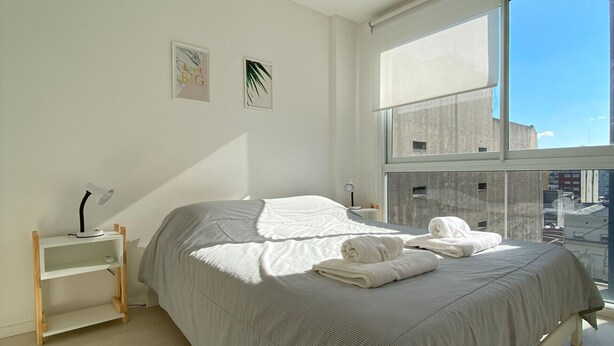 Une chambre avec une grande fenêtre, un lit recouvert d’une couverture à rayures, une table de chevet avec une lampe et une petite étagère.