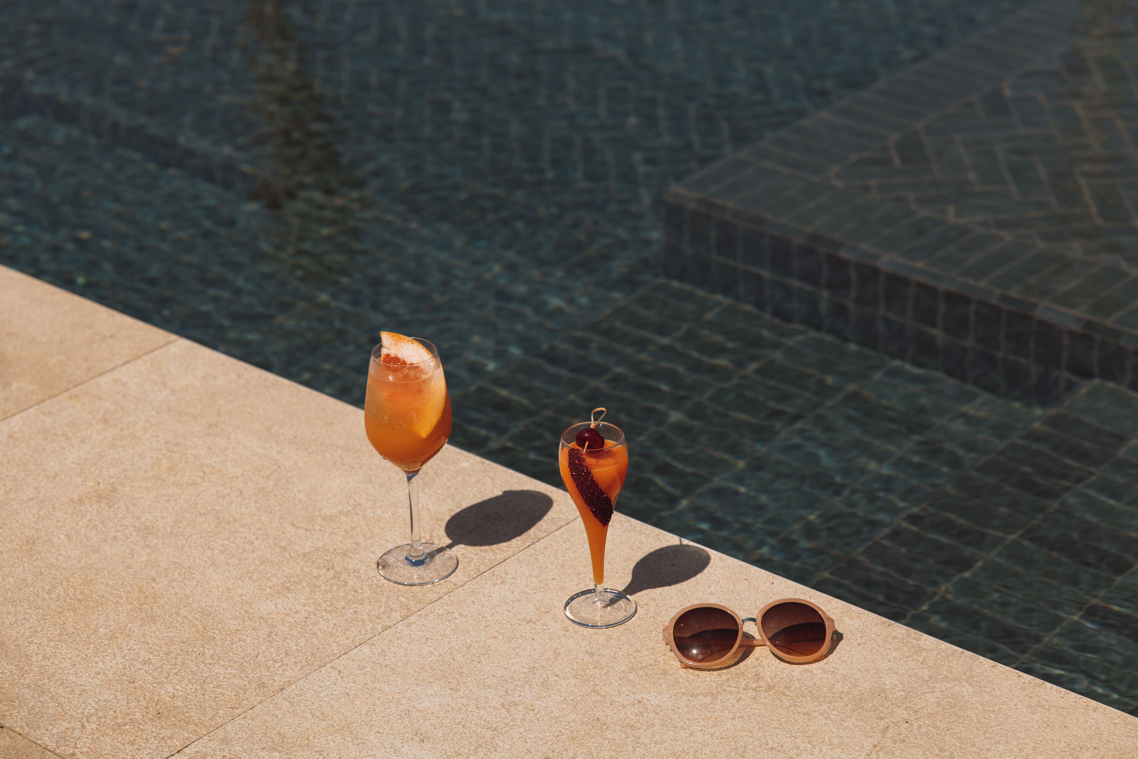 A poolside setting with two drinks and a pair of sunglasses on a tiled surface.