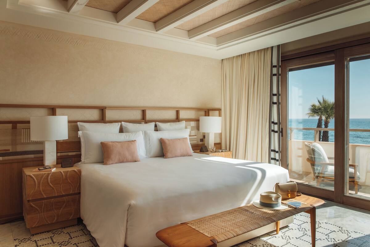 Ein großes Bett mit weißer und beiger Bettwäsche, ein Nachttisch mit Lampe und Meerblick durch ein großes Fenster.