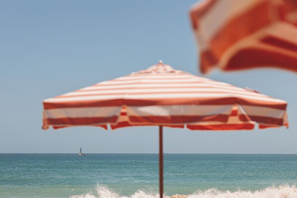 Cena de praia com um guarda-sol listrado e um veleiro ao longe.