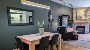 Una sala de estar moderna con comedor, que incluye una mesa de comedor de madera, sillas negras, una chimenea de piedra y un espejo grande en la pared.