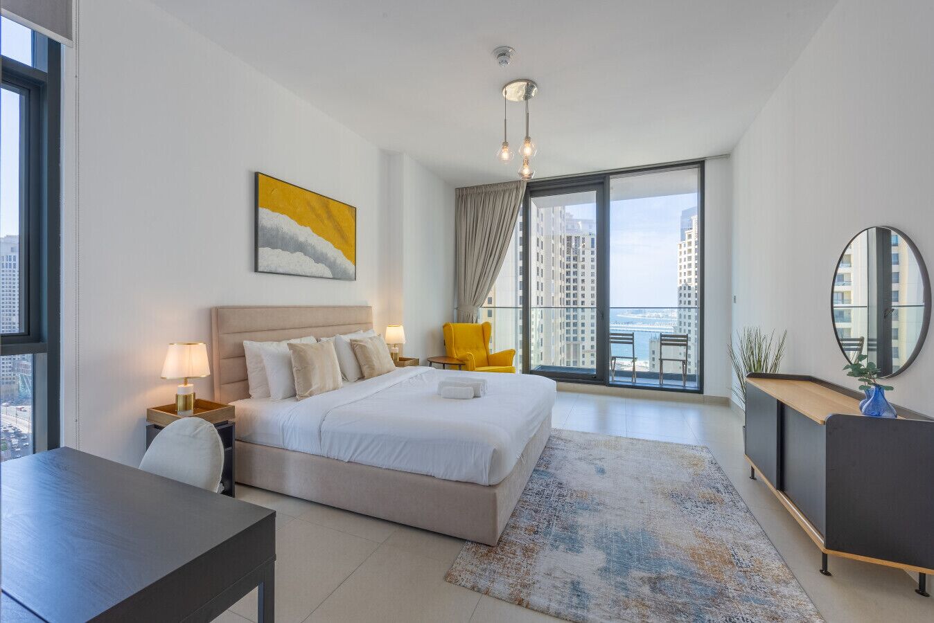 Quarto moderno com uma cama grande, uma escrivaninha, uma cadeira e varanda com vista para a cidade.