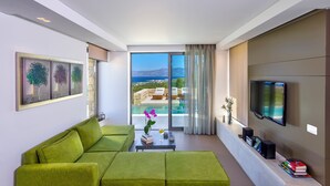 A modern living room with a green sofa, a coffee table, and a TV. There is a view of a pool and mountains in the background.