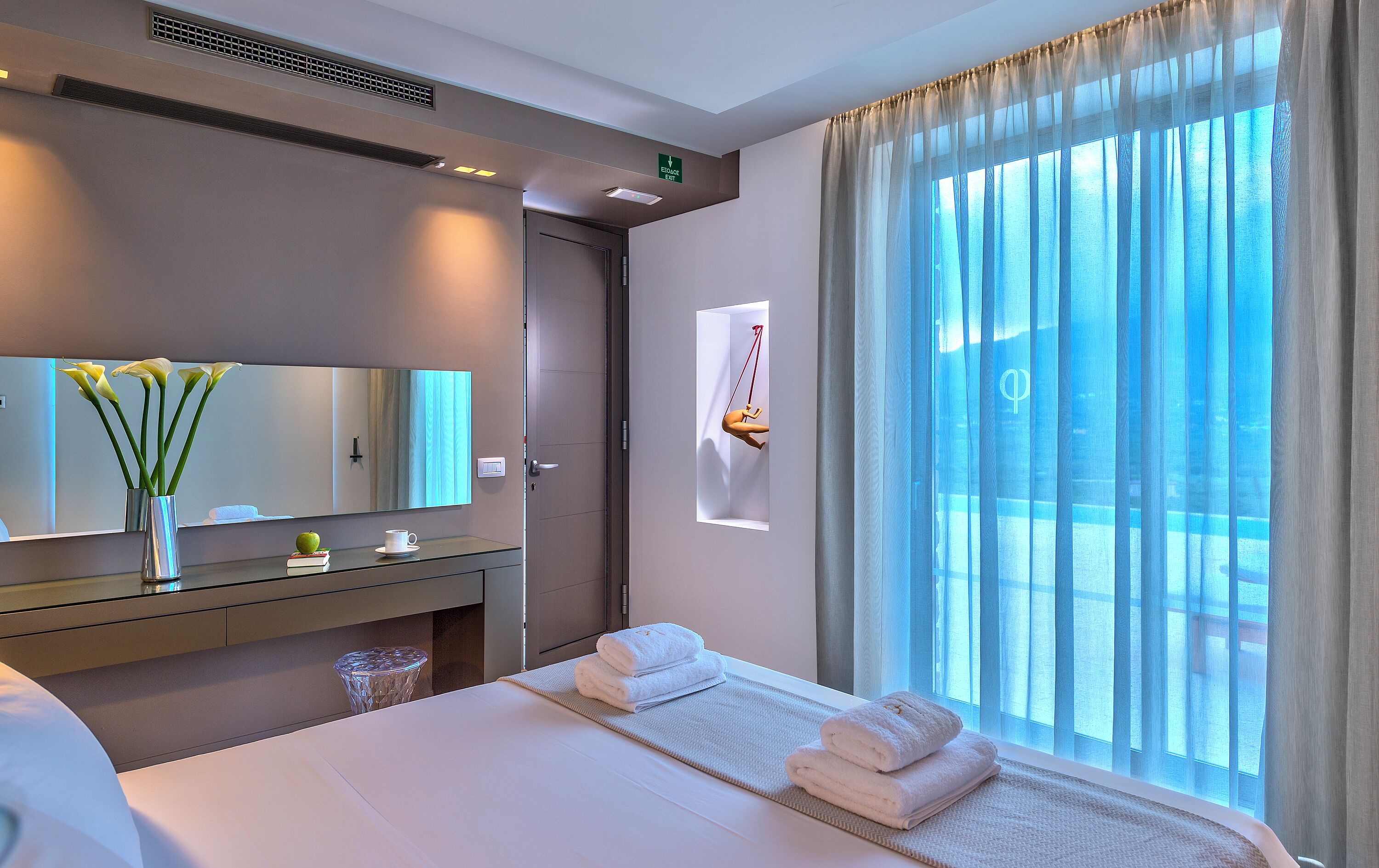 A hotel room with a bed, a vanity with a mirror, a vase with flowers, and a window with curtains.