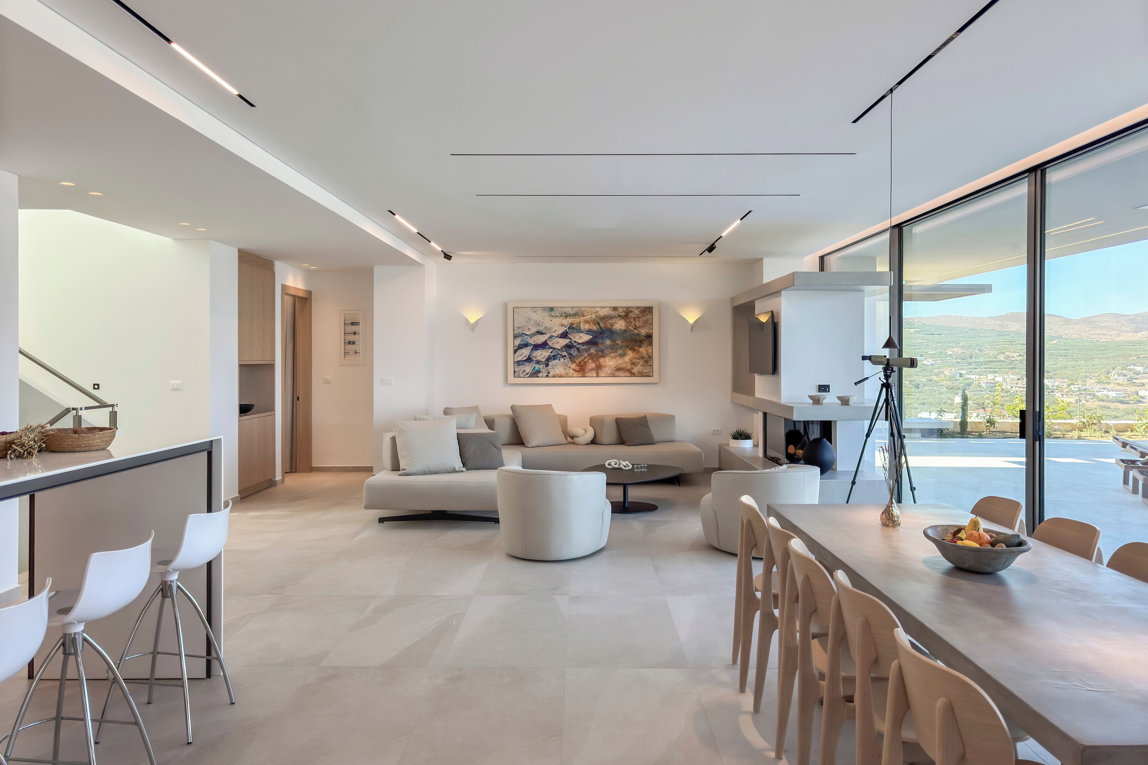 A modern living room with a large window, a sofa, a dining table, and a kitchen area.