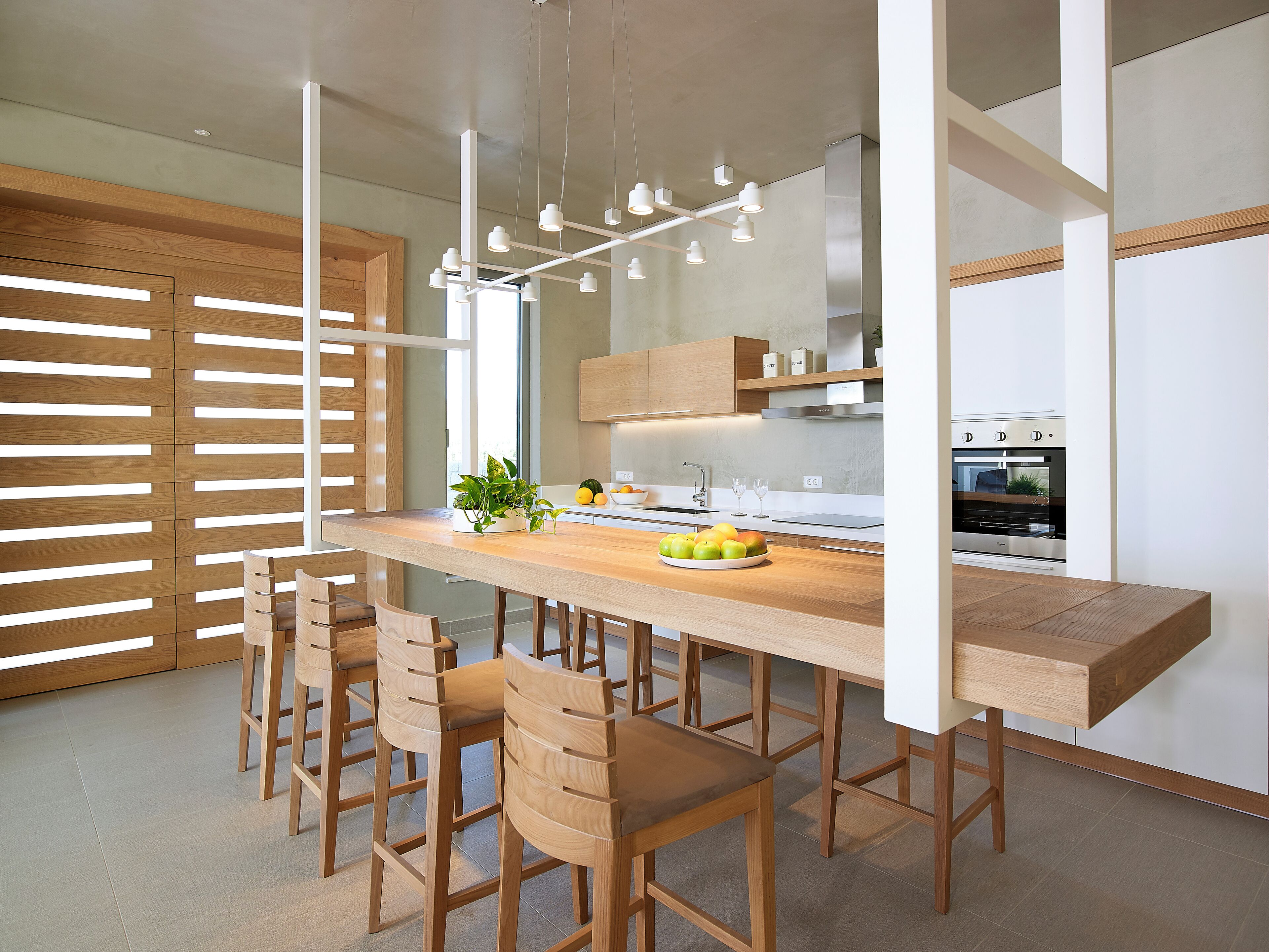 A modern kitchen with a long wooden table, six chairs, a built-in oven, and pendant lighting.